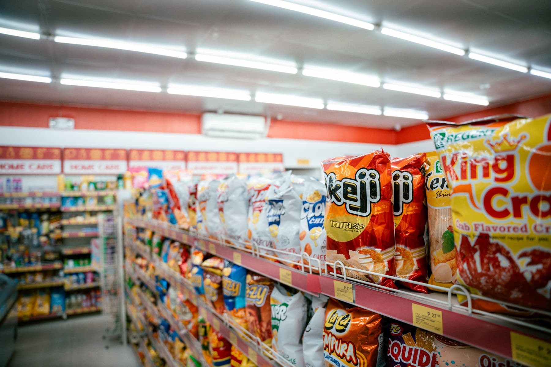A supermarket aisle stacked with snack packs and household lines, shelf labels along the edge