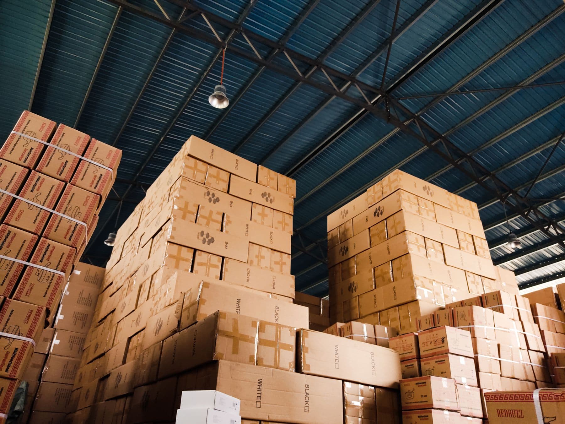 Cartons of packed goods stacked in blocks to the roof of a warehouse, seen from the floor