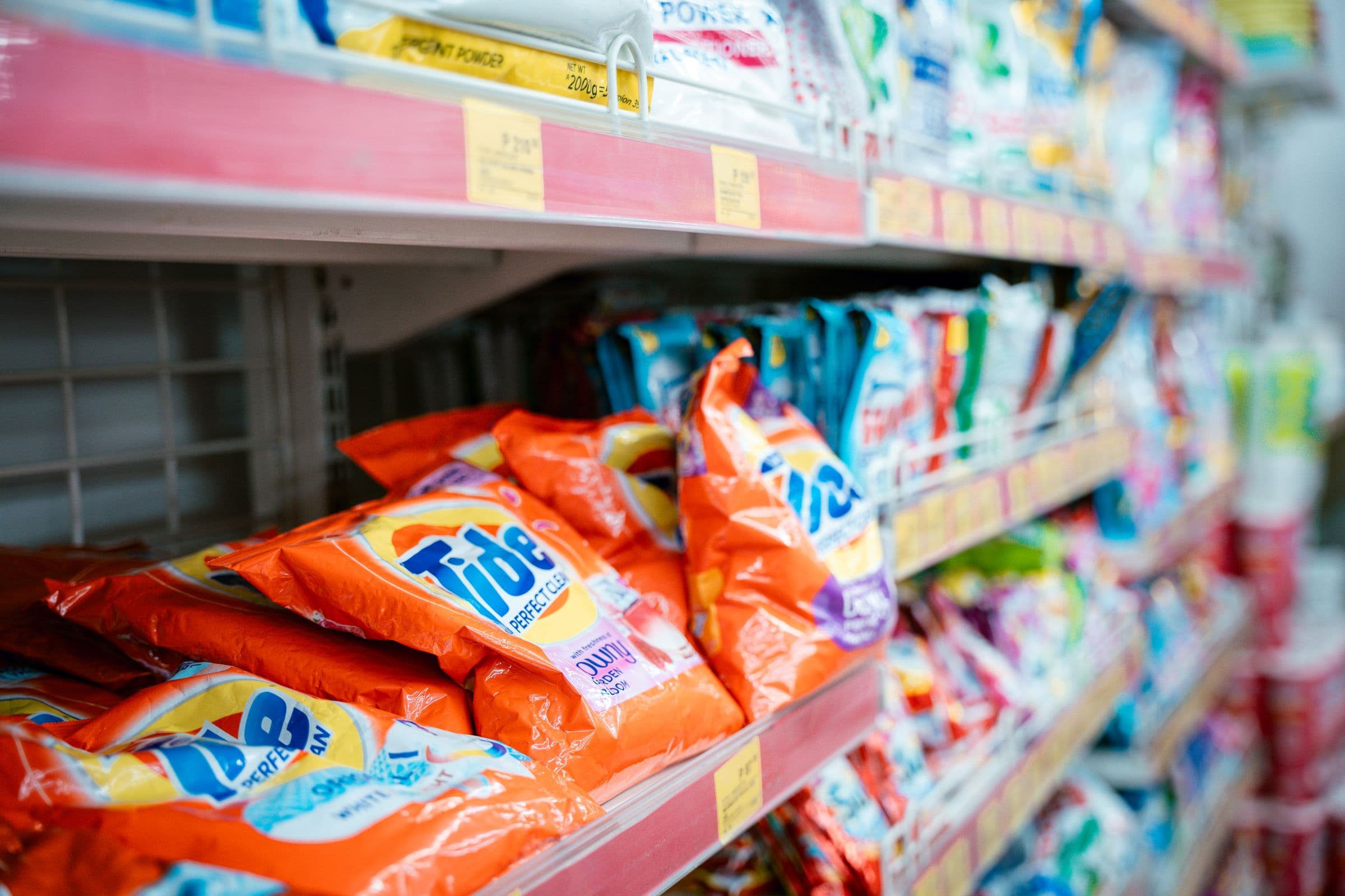 Sachets of laundry detergent racked on a supermarket shelf, price labels running along the shelf edge