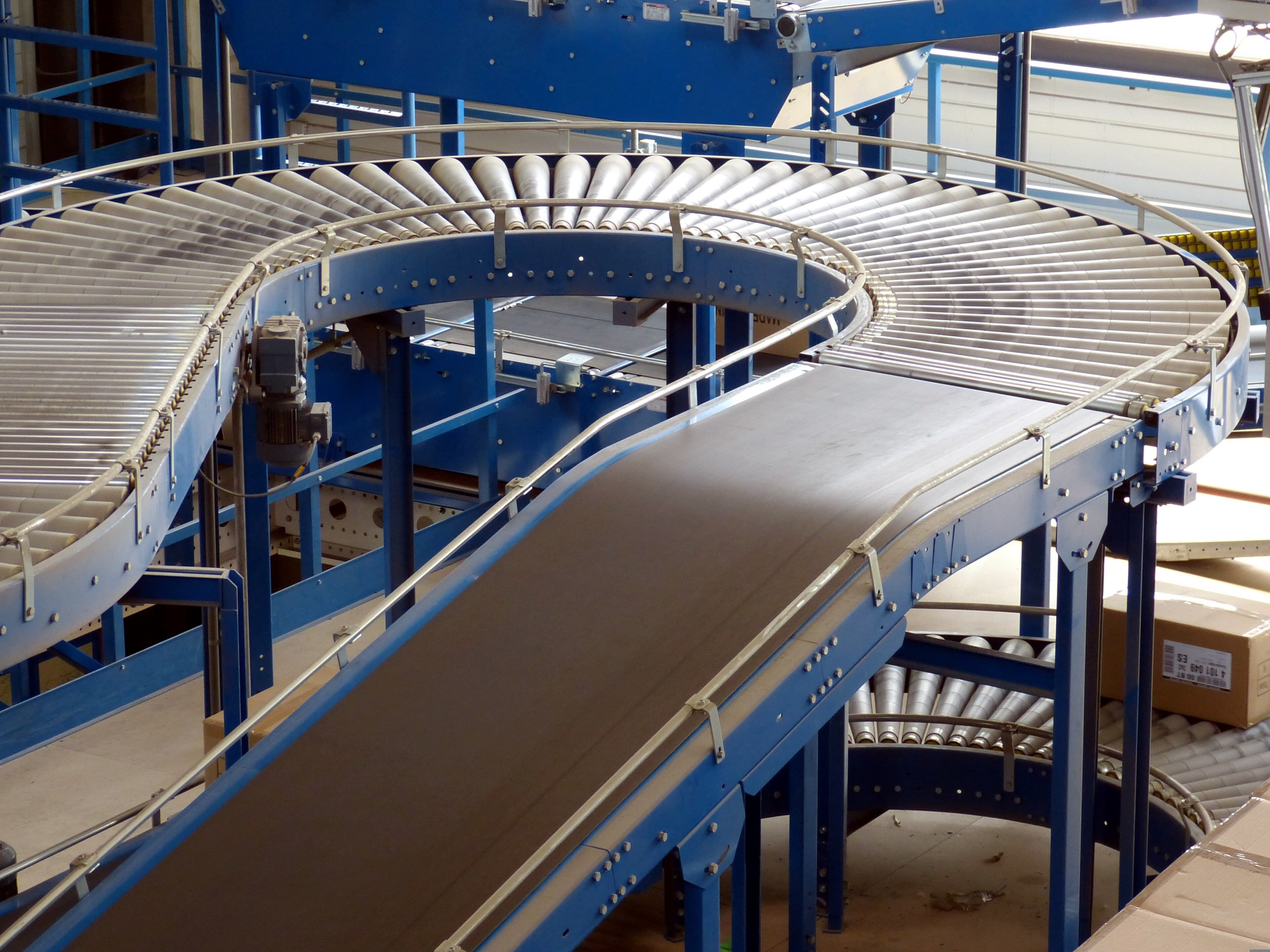 A roller and belt conveyor curving back on itself on blue steel framing inside a warehouse, a labelled carton waiting on the rollers at the right