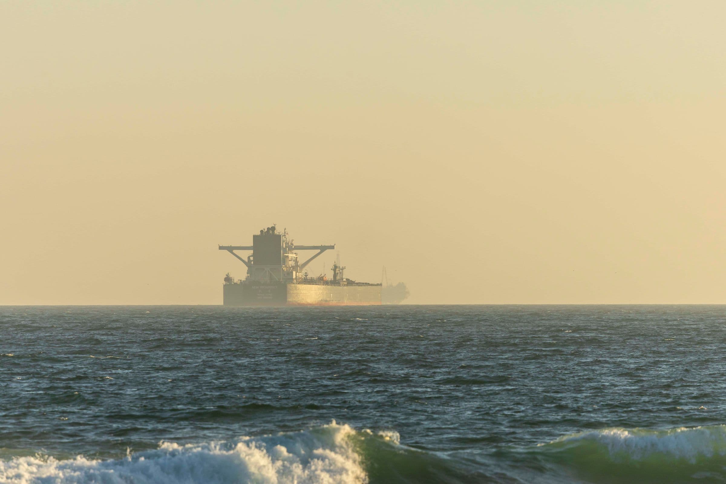 A bulk carrier standing off the coast in the haze of a low sun, seen across breaking surf