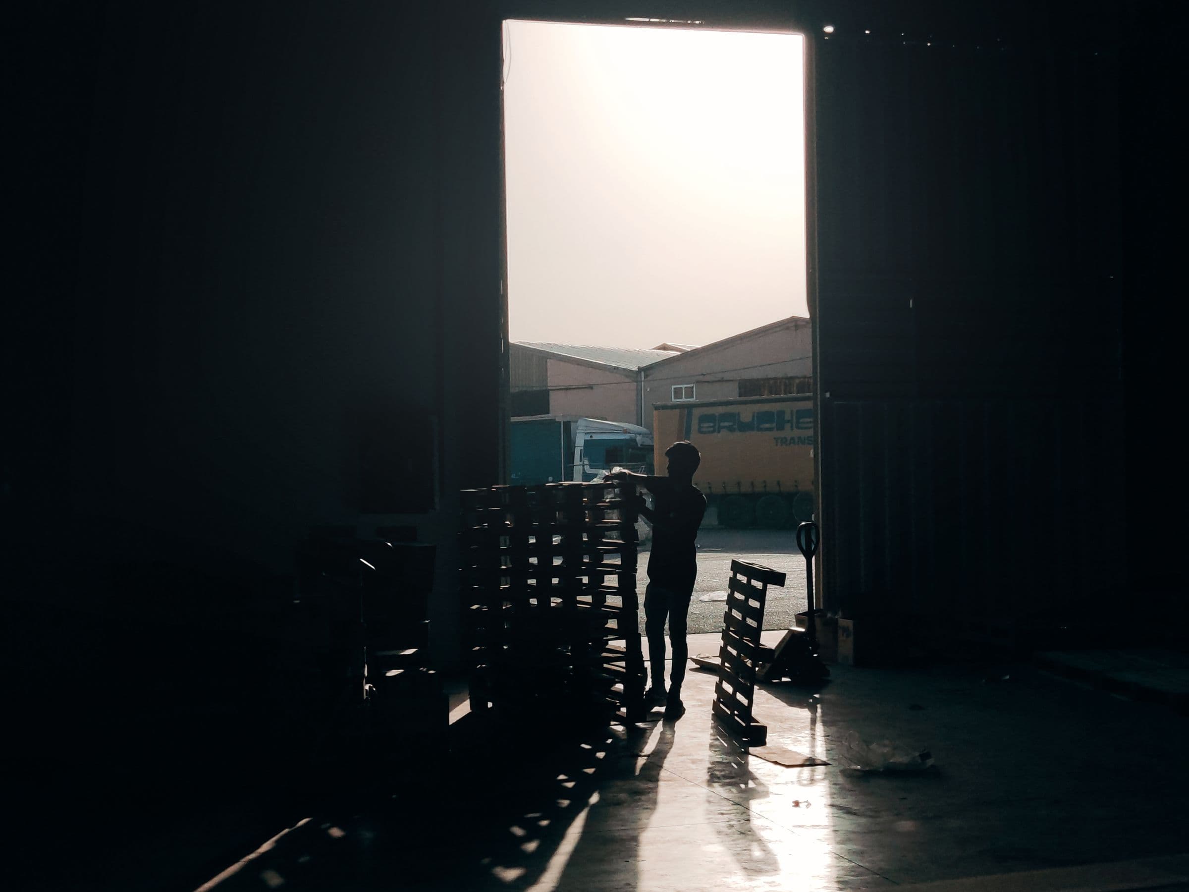 A worker stacking wooden pallets in a warehouse doorway, daylight and a parked trailer behind them