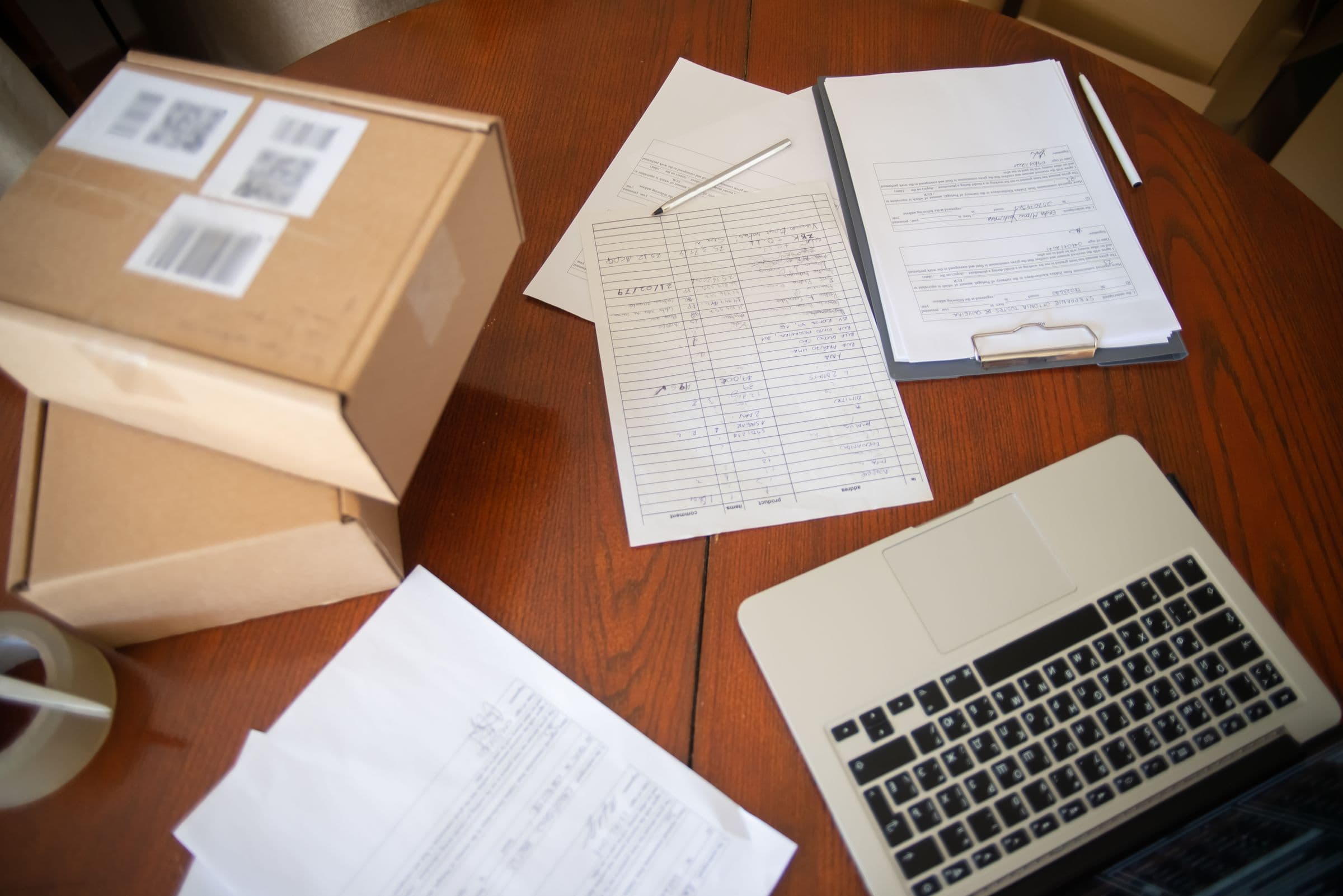 A signed form on a clipboard beside a handwritten packing list, a laptop and two labelled cartons on a desk