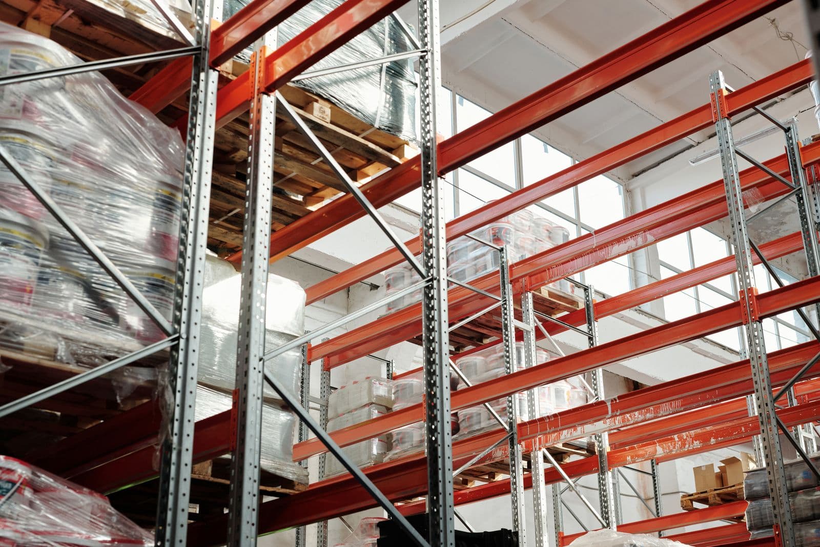 Shrink-wrapped pallets stacked on orange racking in a distribution warehouse, seen from below