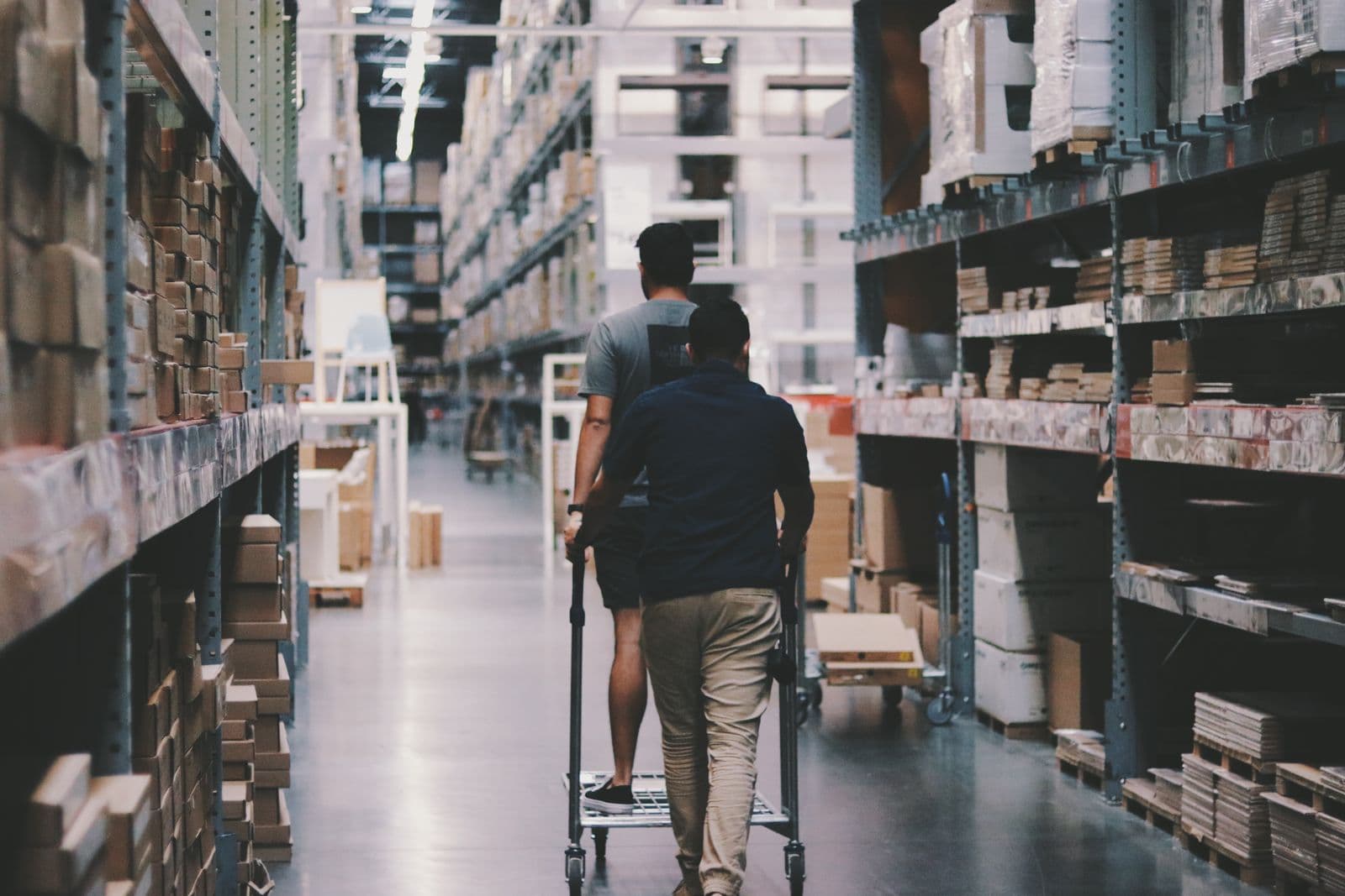 Two people pushing a flatbed trolley down an aisle of racked cartons in a cash-and-carry warehouse