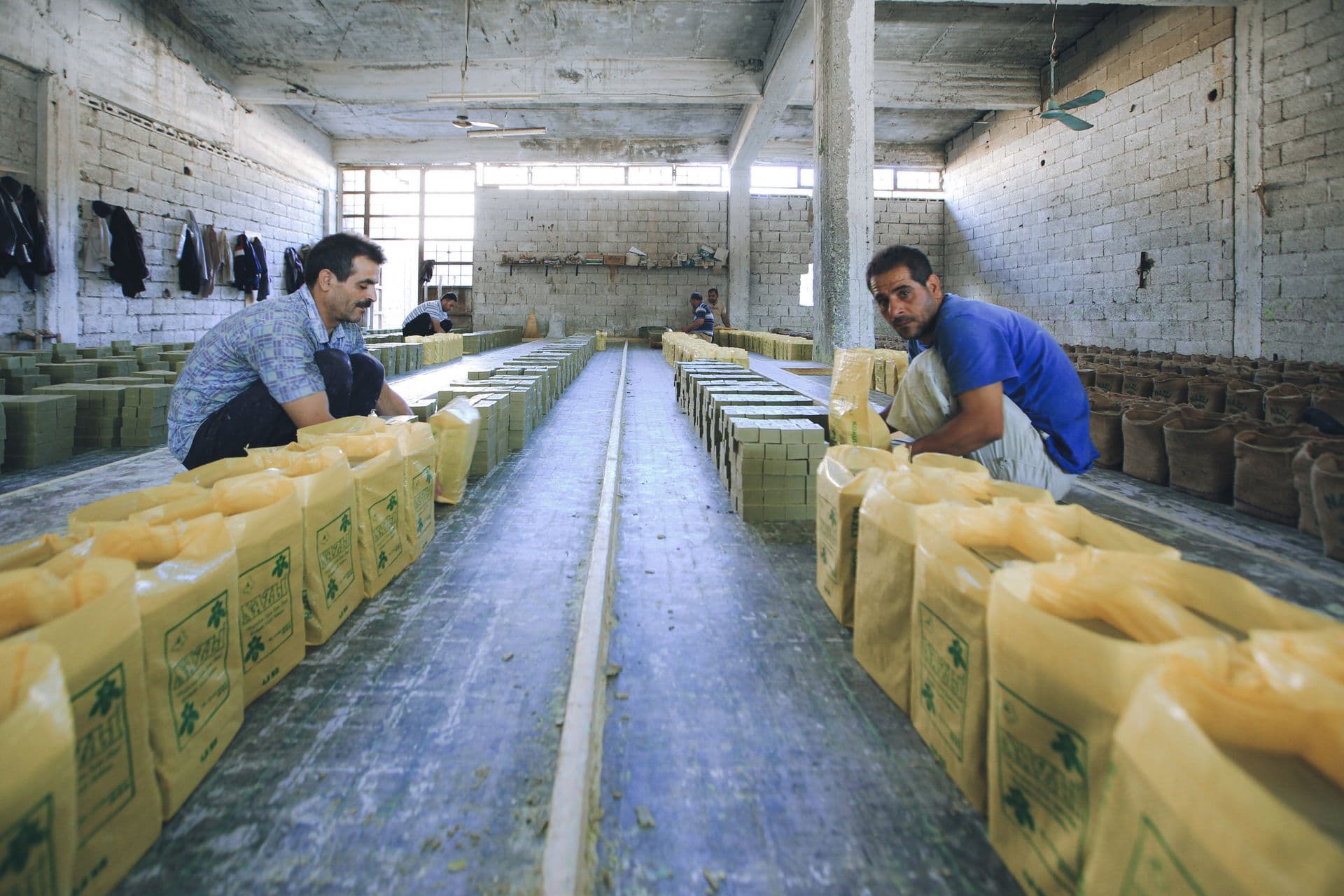 Two men working in a soap factory, crouched near piles of packaged soap inside an industrial hall.