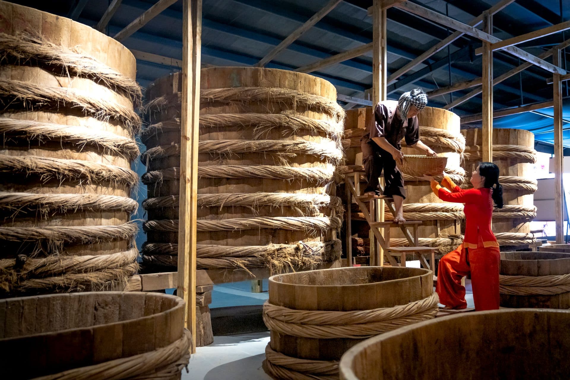 Male worker standing on ladder and giving bowl to female colleague while working in fish sauce factory