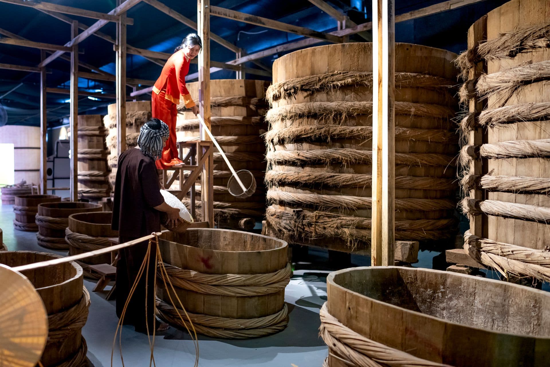 Ethnic female with scoop standing on ladder and preparing fish sauce in wooden container with coworker