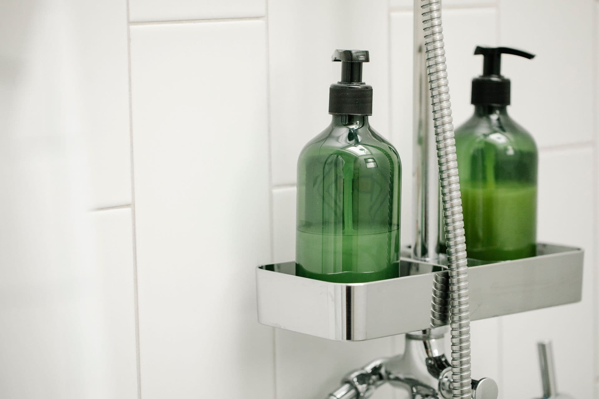 Green dispensers with body lotion on shelf on shower system in bathroom against tiled wall