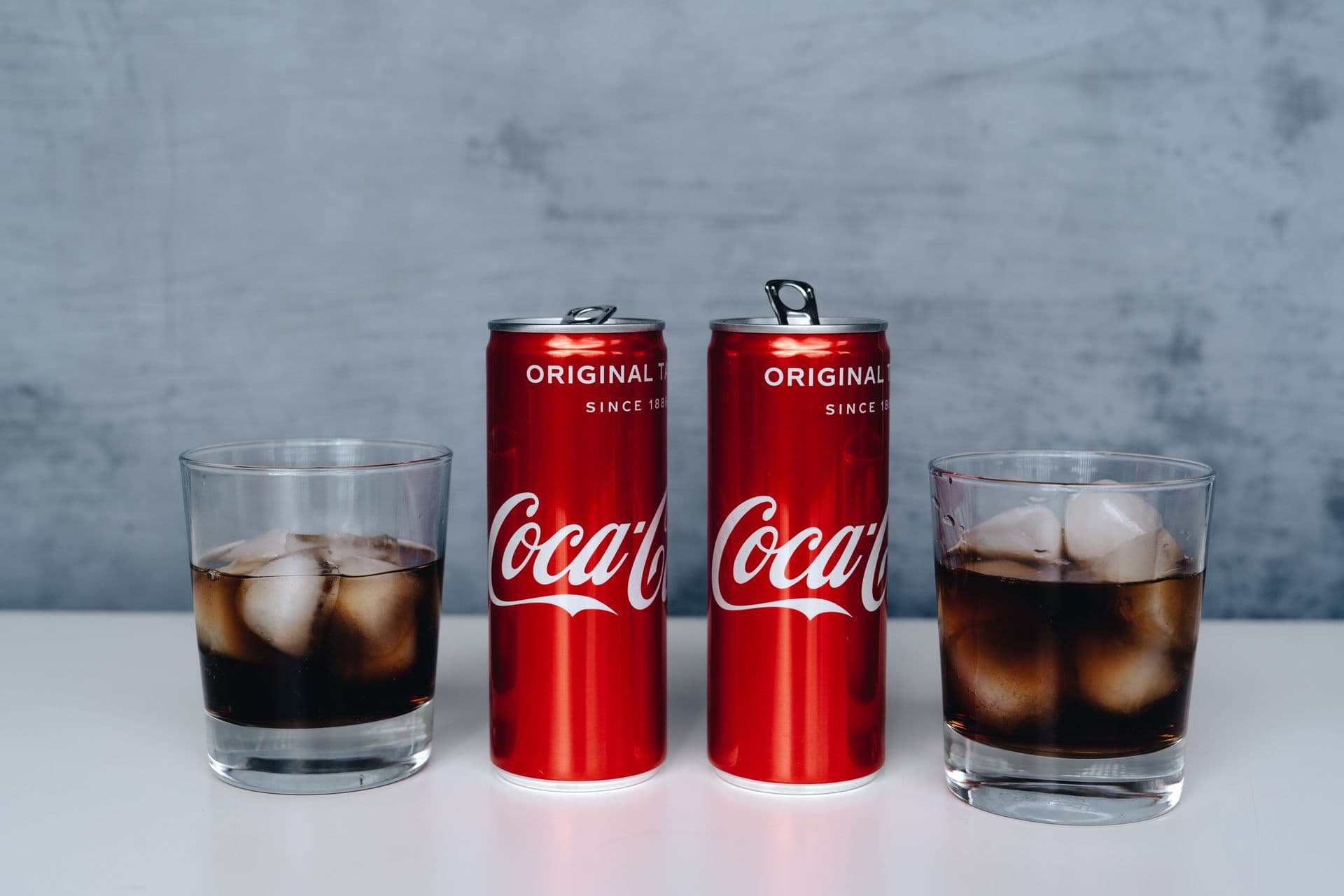 Two cans of soda with ice-filled glasses on a minimalistic background.