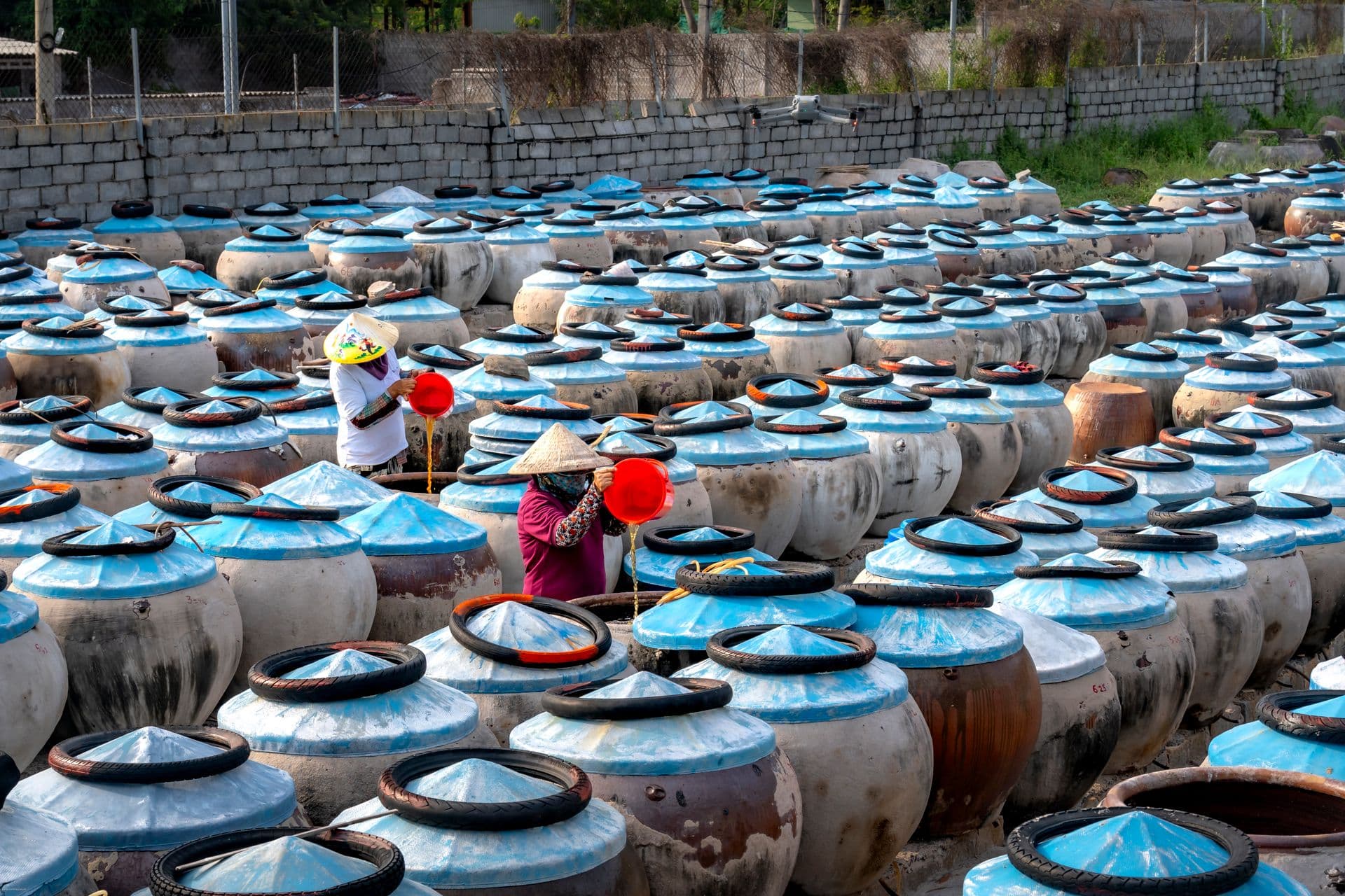 Two workers tend to large fermentation pots in an outdoor market setting, showcasing traditional methods.