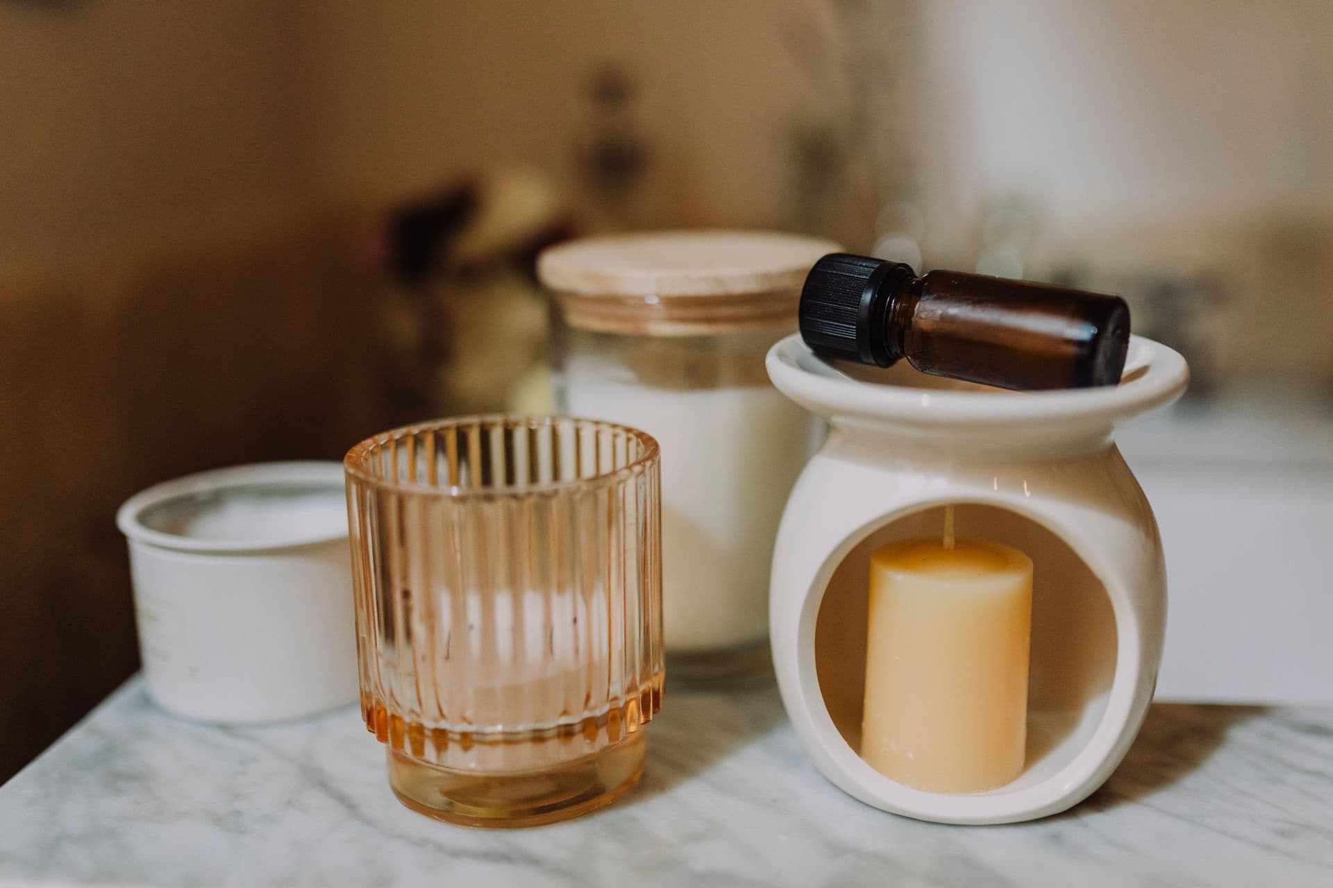 Candles in ceramic and glass jars beside a bottle of essential oil on marble